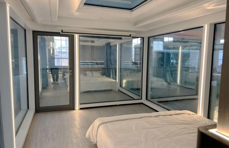 Спальня в LUBAN CABIN K70 с окружающими стеклянными окнами и световым люком, обеспечивающим обильное естественное освещение.